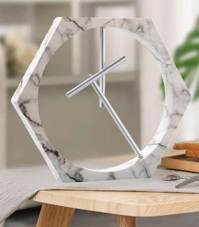 Relógio de mesa com estrutura de mármore branco e ponteiros metálicos, em estilo contemporâneo, colocado numa mesa de madeira com utensílios de cozinha ao fundo.