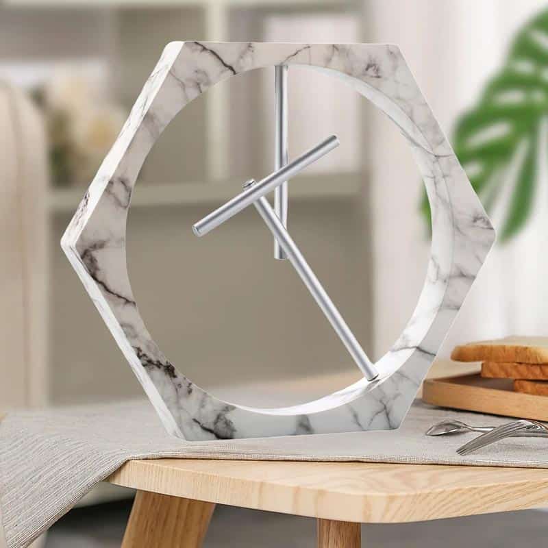 Relógio de Mesa de Design Moderno Relógio de mesa com estrutura de mármore branco e ponteiros metálicos, em estilo contemporâneo, colocado numa mesa de madeira com utensílios de cozinha ao fundo.