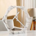 Relógio de parede com estrutura hexagonal de mármore branco e ponteiros metálicos finos, decorativo e moderno.