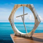 Relógio de areia de mármore com estrutura hexagonal e ponteiro de metal, colocado numa mesa de madeira com vista para o mar azul e céu claro.