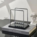 Pêndulo de Newton com cinco bolas de metal suspensas por fios finos, estrutura de metal e base de madeira escura, colocado sobre um livro de ciência.