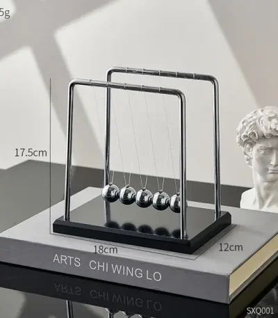 Pêndulo de Newton com cinco bolas de metal suspensas por fios finos, estrutura de metal e base de madeira escura, colocado sobre um livro de ciência.