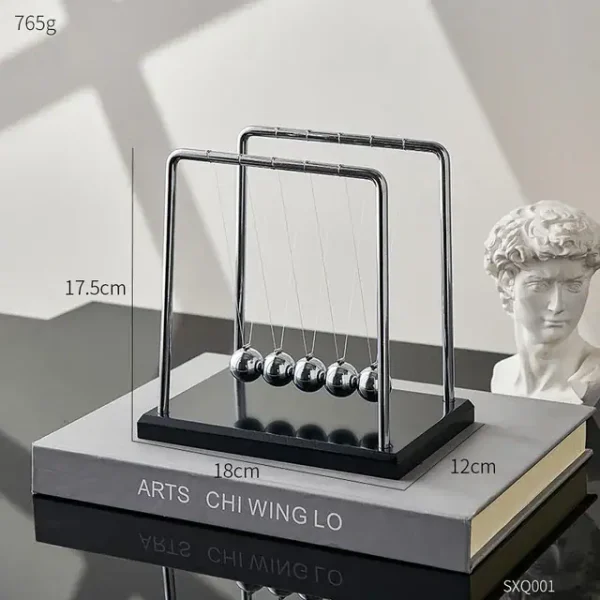 Pêndulo de Newton com cinco bolas de metal suspensas por fios finos, estrutura de metal e base de madeira escura, colocado sobre um livro de ciência.