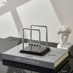 Pêndulo de Newton com bolas de metal suspensas por fios finos, colocado sobre um livro de arte, com uma escultura clássica ao lado, em ambiente bem iluminado.