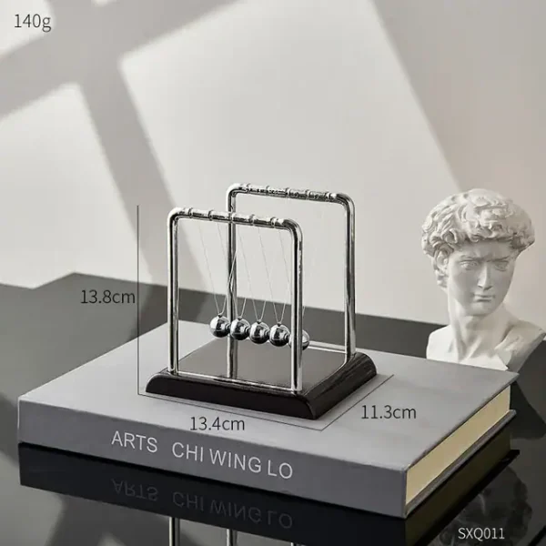 Pêndulo de Newton com bolas de metal suspensas por fios finos, colocado sobre um livro de arte, com uma escultura clássica ao lado, em ambiente bem iluminado.