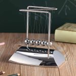 Pêndulo de Newton com bolas de metal suspensas por fios finos, base metálica brilhante, em ambiente de sala de aula ou escritório.