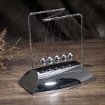 Pêndulo de Newton metálico com cinco bolas suspensas por fios finos, em base de metal brilhante, sobre superfície de madeira escura.