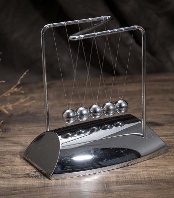 Pêndulo de Newton metálico com cinco bolas suspensas por fios finos, em base de metal brilhante, sobre superfície de madeira escura.