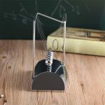 Pêndulo de Newton de metal com bolas suspensas por fios finos, em cima de uma base preta, com quadro negro ao fundo e livro ao lado.