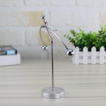 Pêndulo de Newton com bolas de metal suspensas por fios finos, em posição de equilíbrio, sobre uma base circular de metal, com fundo de parede de tijolos brancos e planta verde ao lado.