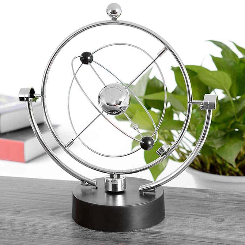 Pêndulo de Newton com esferas de metal suspensas por fios finos, mostrando movimento de balançar, em ambiente de escritório com plantas e livros ao fundo.