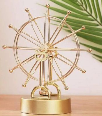 Pêndulo de Newton com esfera de metal dourada, rodas e hastes em disposição radial, sobre base dourada, com folha de palmeira ao fundo.