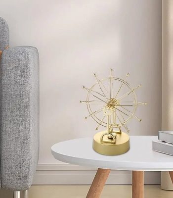 Pêndulo de Newton com estrutura dourada, rodas em movimento e bolas suspensas, colocado numa mesa branca ao lado de livros.