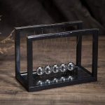 Pêndulo de Newton com bolas metálicas suspensas por fios finos dentro de uma estrutura de metal preta, colocado sobre uma superfície de madeira escura.