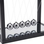 Conjunto de cinco esferas de metal brilhantes suspensas por fios finos, refletindo a estrutura ao redor, com base de vidro e estrutura de metal preto.