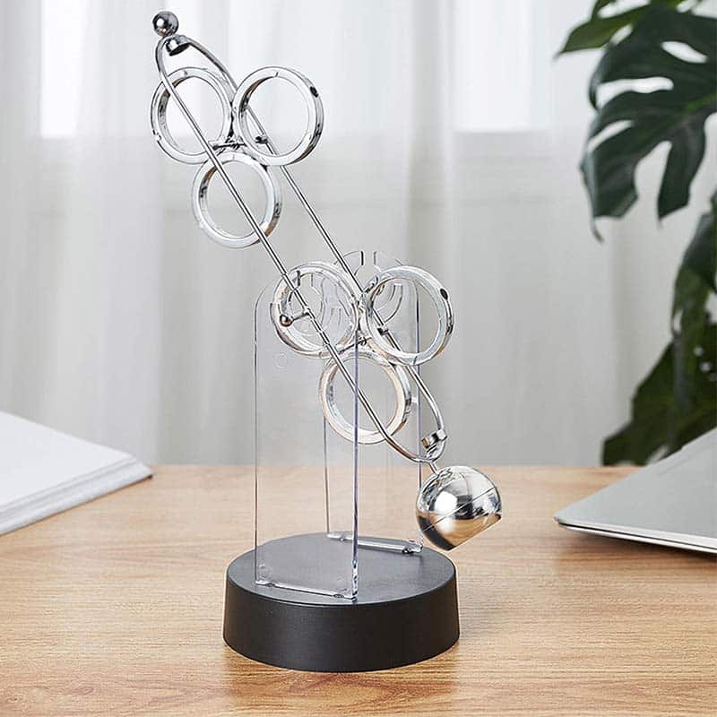 Pêndulo de Newton com várias esferas de metal suspensas por fios finos, em diferentes alturas, sobre base preta, em ambiente de escritório com plantas e luz natural.