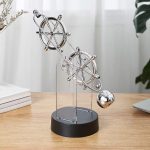 Pêndulo de Newton com duas rodas de metal suspensas por fios finos, base preta e ambiente bem iluminado com plantas ao fundo.