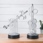 Dois pêndulos de Newton de metal e acrílico, um com várias bolas e outro com círculos, em exibição sobre uma mesa de madeira com fundo branco.