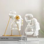 Relógio de areia com estrutura metálica dourada e figura de astronauta de pelúcia a segurar o vidro, decorativo e educativo.