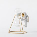 Miniatura de astronauta em traje espacial segurando uma ampulheta com areia branca, decorativa e educativa.