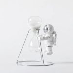 Miniatura de astronauta branco com capacete prateado segurando uma ampulheta de vidro com areia branca, sobre suporte metálico.