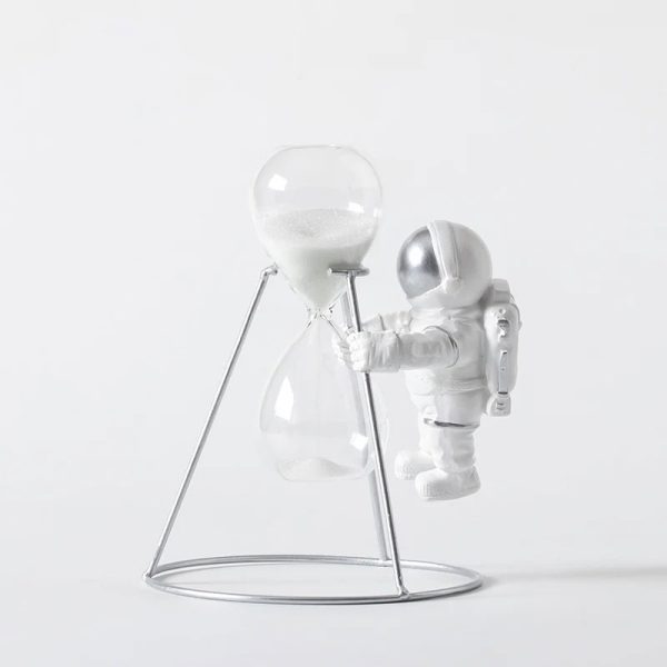 Miniatura de astronauta branco com capacete prateado segurando uma ampulheta de vidro com areia branca, sobre suporte metálico.