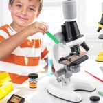 Criança a usar um microscópio com tubo de luz, pipeta e amostras, em ambiente de laboratório educativo.