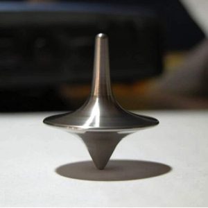 Pião de metal com ponta afiada e corpo arredondado, em rotação sobre uma superfície plana, criando sombra e efeito de movimento.