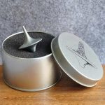 Pião metálico com design futurista, colocado numa caixa de metal com tampa, vista de cima e de lado, com fundo de parede cinzenta.