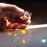 Prisma de cristal transparente segurado por uma mão, dispersando luz que cria reflexos coloridos no plano.