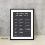 Quadro com tabela do Código Morse, mostrando letras e números em pontos e traços, em fundo preto e letras brancas, apoiado numa parede de cimento.