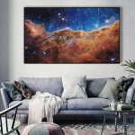 Painel de parede com imagem detalhada da Nebulosa da Quilha, mostrando cores vibrantes e estrelas brilhantes, decorando uma sala moderna com sofá cinza e plantas.