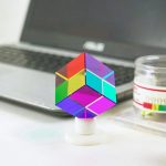Prisma cúbico colorido com faces refletoras, colocado numa mesa junto a um portátil e uma caixa de pequenos objetos.