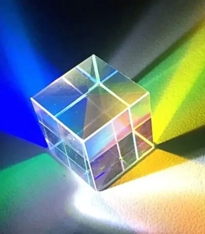 Cubo de vidro transparente com prismas internos que dispersam a luz em cores do espectro visível, projetando um arco-íris de luz na superfície.
