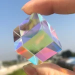 Pequeno prisma de vidro em forma de cubo, refletindo cores do arco-íris, segurado entre os dedos contra o céu claro.