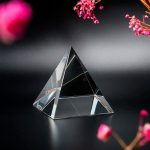 Prisma de vidro transparente em forma de pirâmide, refletindo luz e cores, com flores rosa ao redor em fundo escuro.