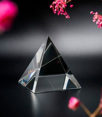Prisma de vidro transparente em forma de pirâmide, refletindo luz e cores, com flores rosa ao redor em fundo escuro.
