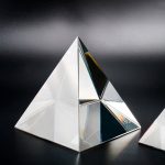 Prisma óptico de vidro em forma de pirâmide com superfícies refletoras e prismáticas, sobre fundo escuro, destacando-se pela transparência e brilho.
