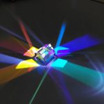 Prisma pentagonal de vidro transparente com luzes coloridas refletidas e dispersas em diferentes direções sobre fundo escuro.