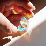 Prisma óptico pentagonal transparente refletindo luz com cores vibrantes, segurado por uma mão com luz natural ao fundo.