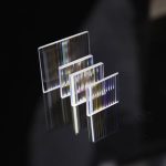 Conjunto de quatro prismas retangulares dicroicos de vidro transparente, com diferentes ângulos e cores refletidas, sobre fundo preto.