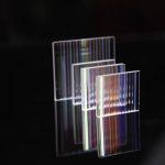 Três prismas retangulares dicroicos transparentes com diferentes cores refletidas, sobre fundo preto, mostrando diferentes ângulos e detalhes de reflexão.