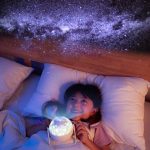 Menina deitada na cama a olhar para projeção de estrelas no teto, segurando uma lanterna de luz suave.