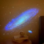 Projeção de uma galáxia azul e amarela no teto, com estrelas dispersas ao redor, criada por um projetor estelar.