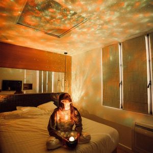 Projetor de estrelas a projetar luzes coloridas no teto e paredes de um quarto escuro, com uma mulher sentada na cama a observar as luzes.