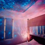 Projeção de estrelas e galáxias no teto de um quarto, criando efeito de céu estrelado com luzes azuis, rosas e brancas.