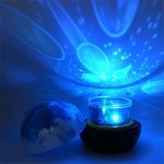 Projetor de galáxia em funcionamento com projeções de estrelas e planetas no teto, luz azul brilhante, com tampa removida ao lado.