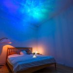 Céu estrelado projetado no teto de um quarto com cama de casal, iluminação suave e decoração temática de espaço.
