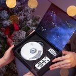 Projetor LED com tema de galáxia, caixa aberta mostrando o dispositivo e acessórios, fundo com árvore de Natal decorada com luzes douradas.