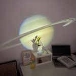 Projeção de Saturno com anéis visíveis, refletida na parede, criando sombra de uma planta e objetos na mesa.